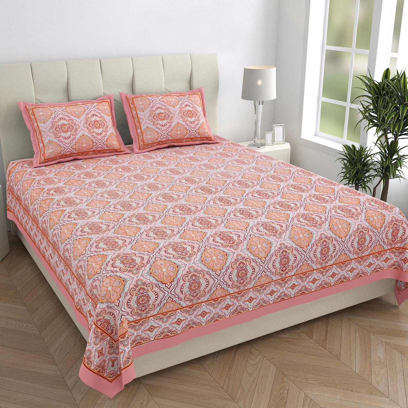 Bed with pink and orange patterned bedspread and pillows in a room with a lamp and plant.