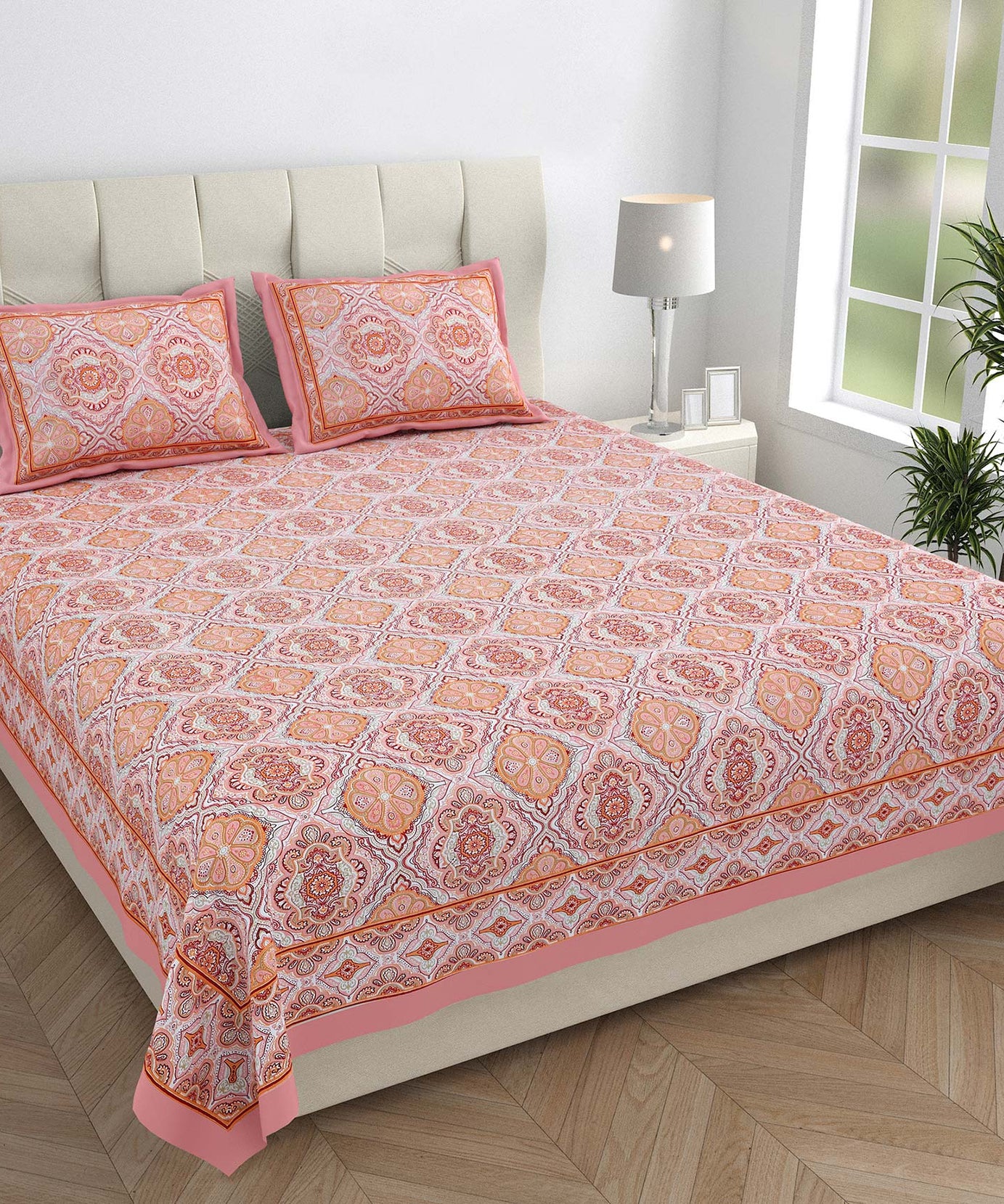 Bed with pink and orange patterned bedspread and pillows in a room with a lamp and plant.