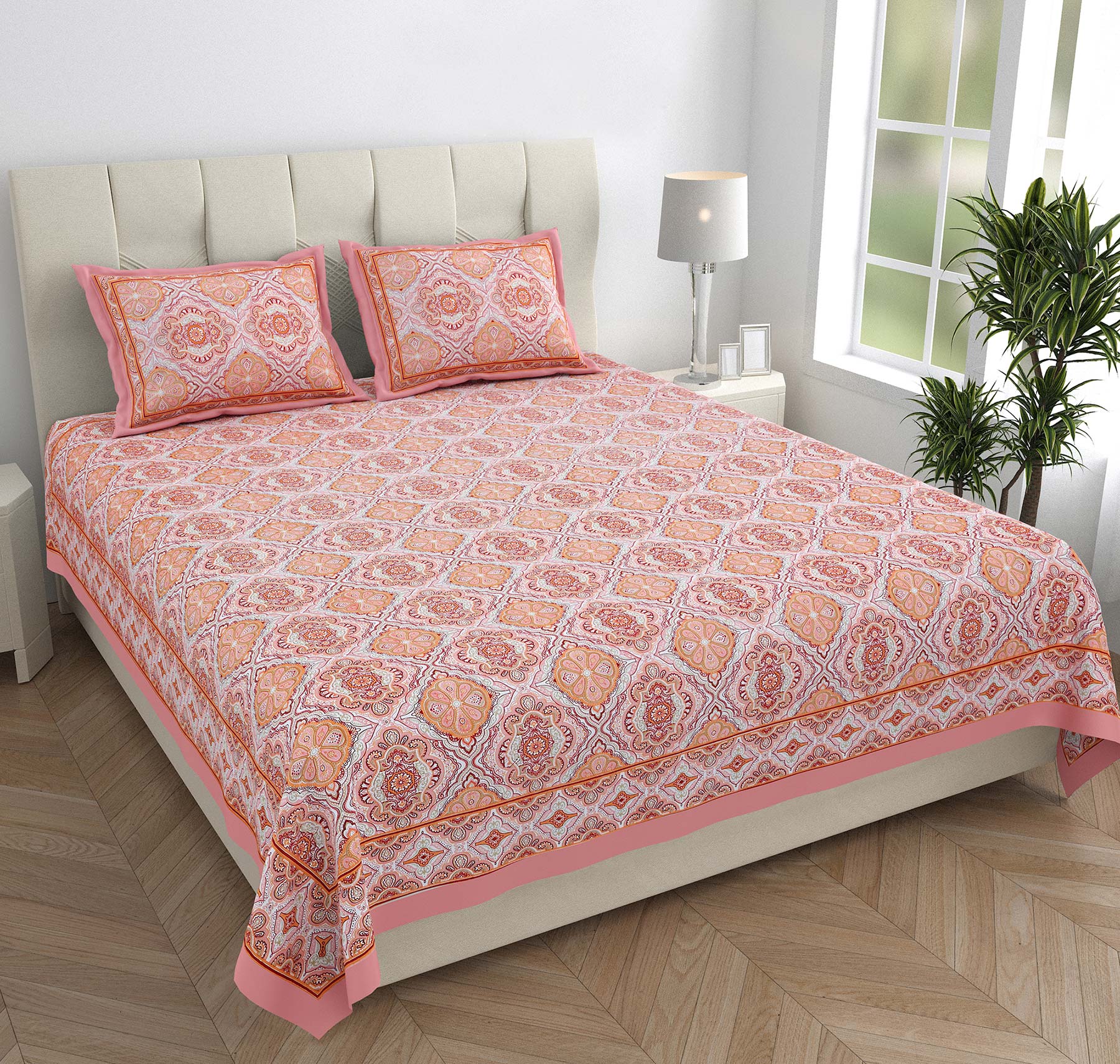 Bed with pink and orange patterned bedspread and pillows in a room with a lamp and plant.